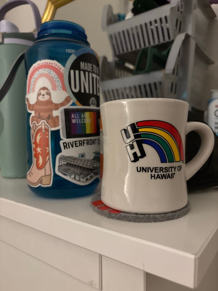 A rainbow University of Hawaii mug next to a blue water bottle covered in stickers 