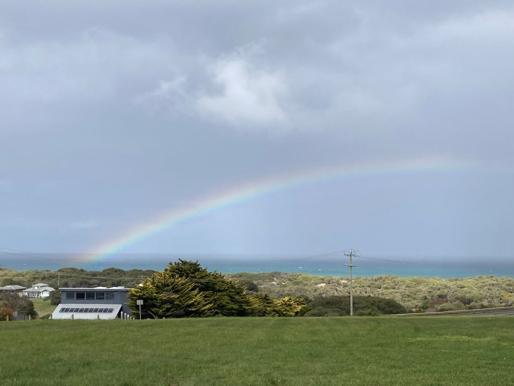 Looking south to Bass Strait. In the foreground an expanse of green lawn dips into natural scrub and dunes. The ocean is steely grey, matching the rain clouds that blanket the horizon. A rainbow glows gently, seeming to rise from one of the posh houses visible among the bushes. Photo by me.