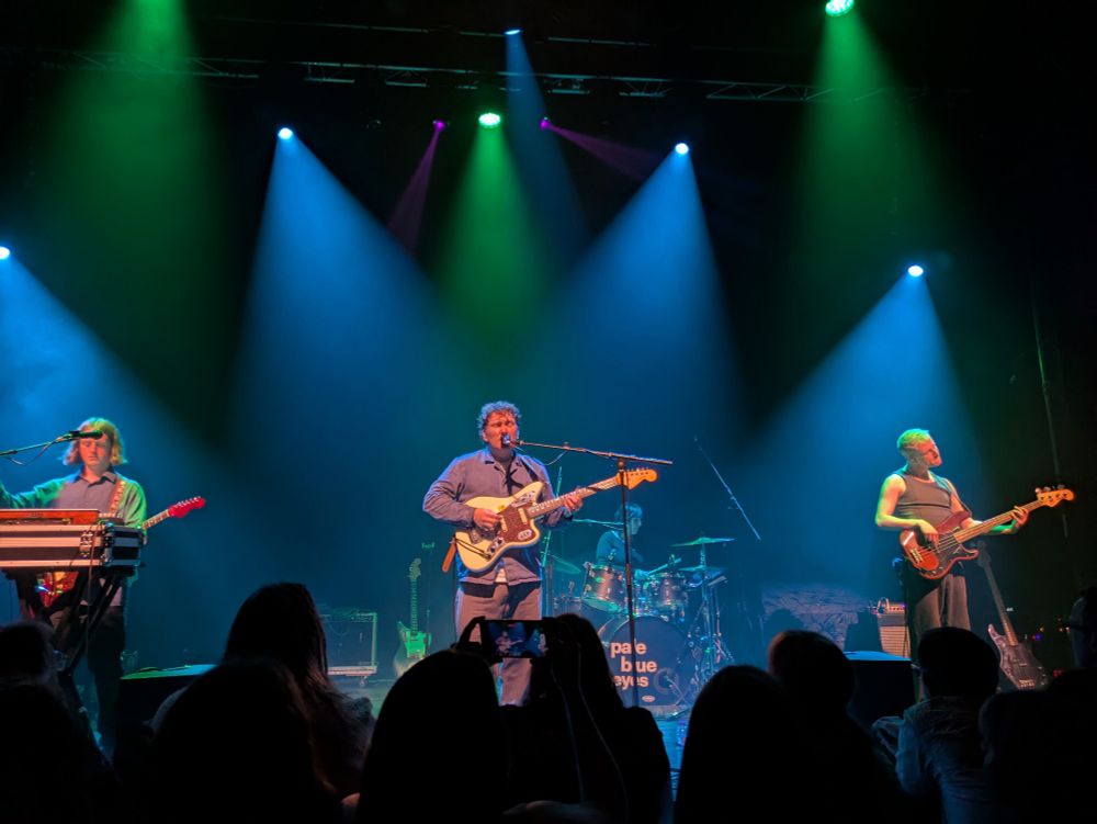 Members of pop group Pale Blue Eyes on stage at Exeter Phoenix, two guitarists, bassist and drummer, bathed in green and blue lights.