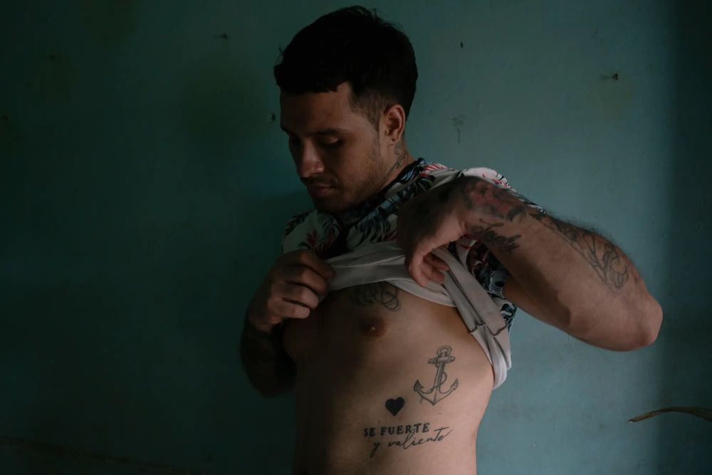 Albert Rodríguez Parra lifts his shirt to display some of his tattoos, including an anchor and a heart with the message Se Fuerte y valiente.