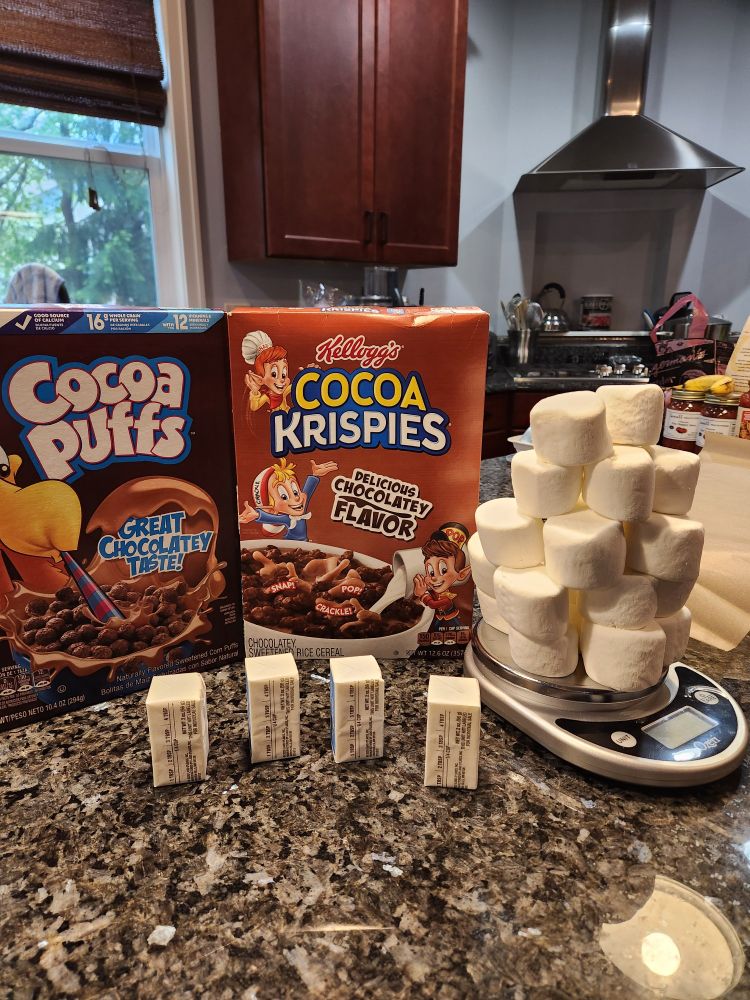 Cocoa Puffs, Cocoa Krispies, and four half-sticks of butter carefully lined up on the counter in preparation for Rice Krispy treat making, next to a kitchen scale with the *exact* number of marshmallows needed weighed out.