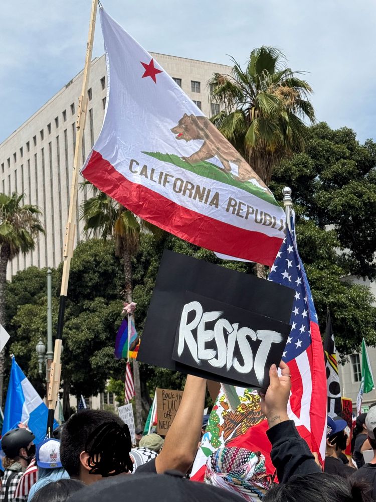 At a protest, with the American flag and California state flag flying, and a sign that says “resist”