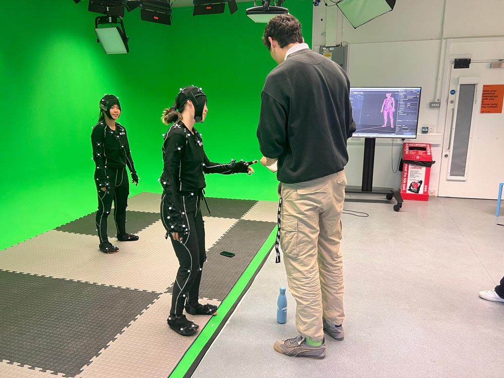 A tall man in jumper and pale trousers writes in a notebook. Two female dancers in motion capture suits stand in a green screen space.