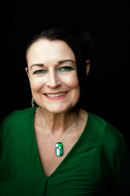 Photograph of Ghislaine Boddington, a white woman with dark hair pulled back off her face. She is wearing blue eyeshadow, a dark green v-neck top and a a bold statement necklace