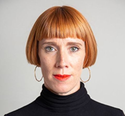 A photo of Lucy Bayliss, a white woman with short cropped red hair, wearing a black polo neck top, gold hoop earrings and red lipstick