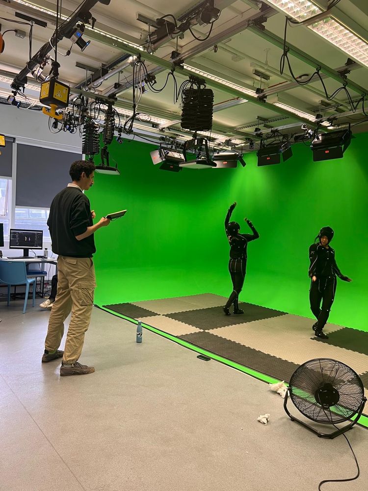 A tall man in jumper and pale trousers writes in a notebook. Two female dancers in motion capture suits stand in a green screen space.