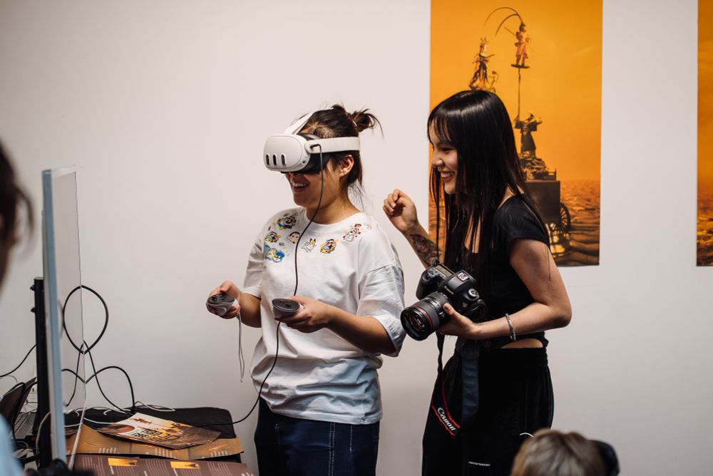 Two young women experimenting with a VR installation, one wearing a headset the other at her side watching the screen