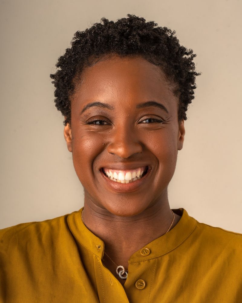 Photograph of Dr Tia-Monique Uzor, a Black woman with short hair wearing a mustard yellow top and smiling for the camera