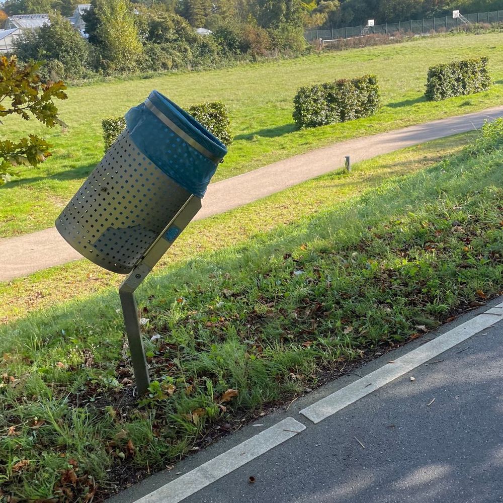 This picture shows a trash can at the side of a street for bicycles. The trash can is placed at a slight angle so that the opening faces passing cyclists. So it's easier for them to put their trash in it without needing to stop cycling.