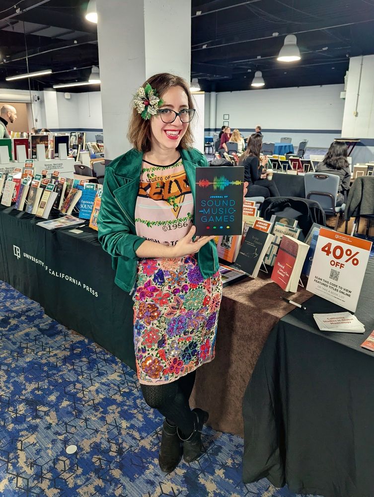 Dana in a multicolored floral pencil skirt, a shirt with the title screen from the original Legend of Zelda game, and a green velvet blazer, holding up a copy of the Journal of Sound and Music in Games