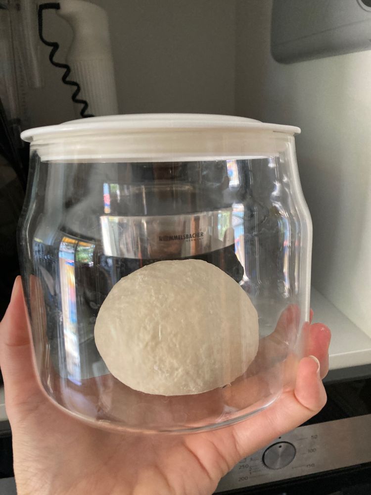 Teigkugel in Glas mit weissem Kunststoffdeckel wird von einer Hand ins Bild gehalten 