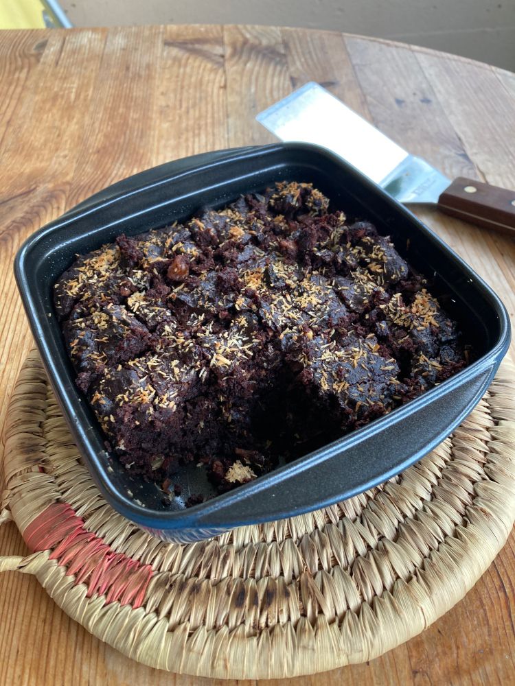 Kleine Form mit Brownies geschnitten mit gebräuntem Kokosraspel obendrauf. Es steht auf einem Bastuntersetzer und dieser auf einem Holztisch