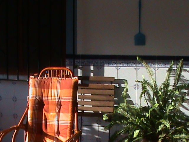 Fotografia en la que se ve una silla/mecedora naranja, detrás la pared de una casa desde afurera con ladrillos de blanco y azul y una ventana. Al lado derecho se asoma una planta y arriba cuelga un matamosquitos de la pared.