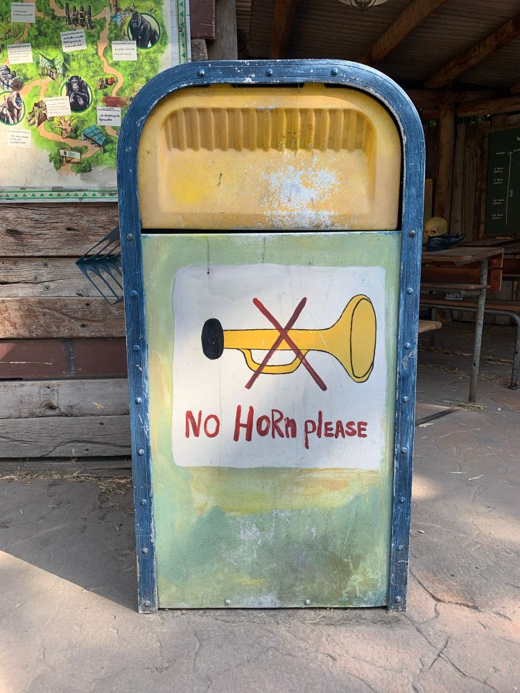 A photo of a trash can in the Ape Mountain area at the Hannover Zoo, with a sign painted in it of a trumpet with a big red X going through it and text underneath that says „no horn please.“