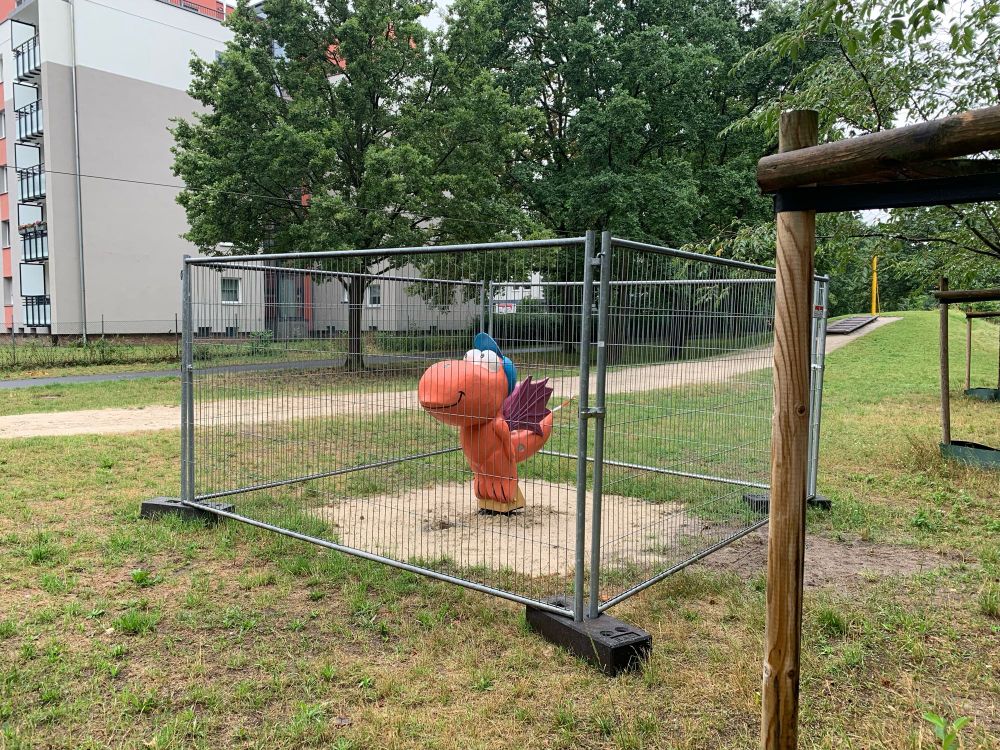 A playground figure of German children‘s book character Coconut the Dragon enclosed in a chain link fence while it’s still under construction. The dragon has a hopeful smile on his face despite being imprisoned.