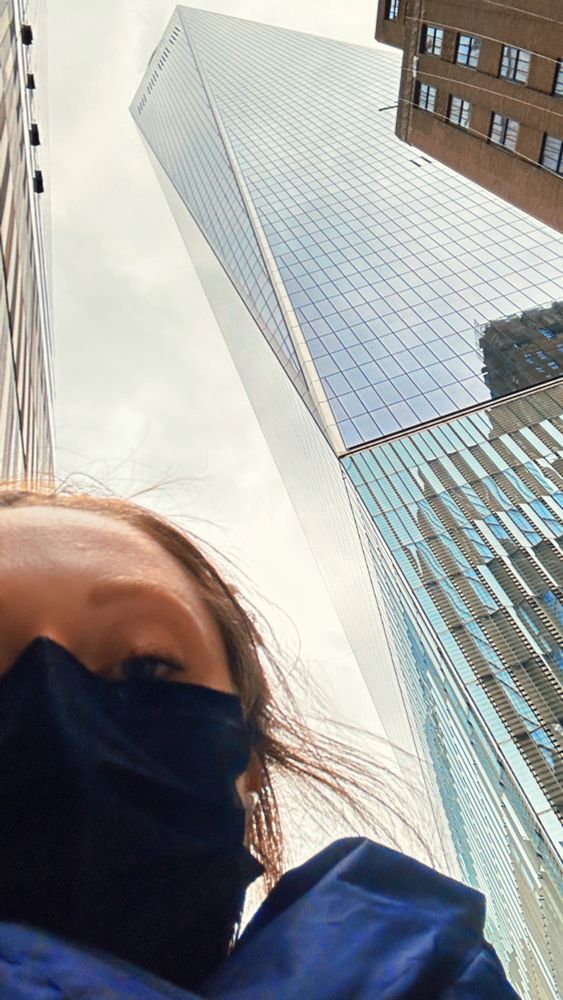 A masked face looks down at the phone screen with 1WTC towering behind her. 