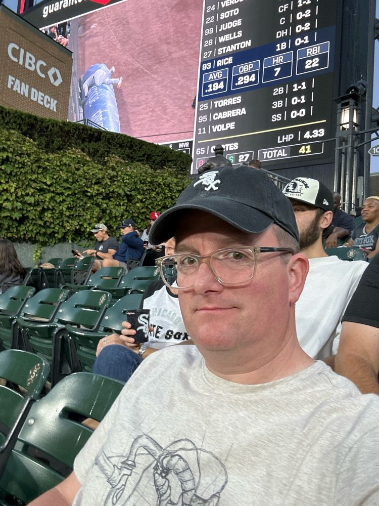 Bespectacled man in a White Sox hat sitting in the outfield of a ballpark