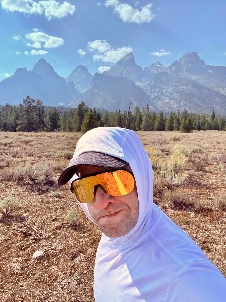 Me, wearing a white sun hoodie, black cap, and reflective sunglasses running along an open field with sagebrush. Pine trees and the Teton Range are visible in the background.