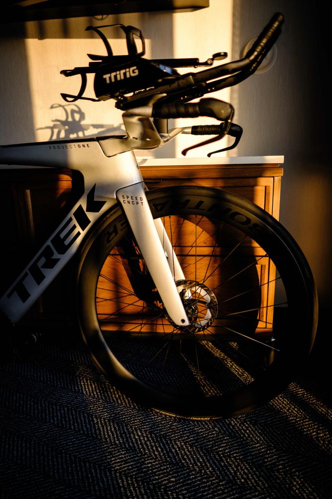 Close-up of the front section of a silver Trek Speed Concept bicycle, featuring aerobars with TriRig arm scoops, deep-section Bontrager wheel, and disc brakes. The bike is positioned indoors with golden morning sunlight casting shadows on the wall and a wooden cabinet in the background.