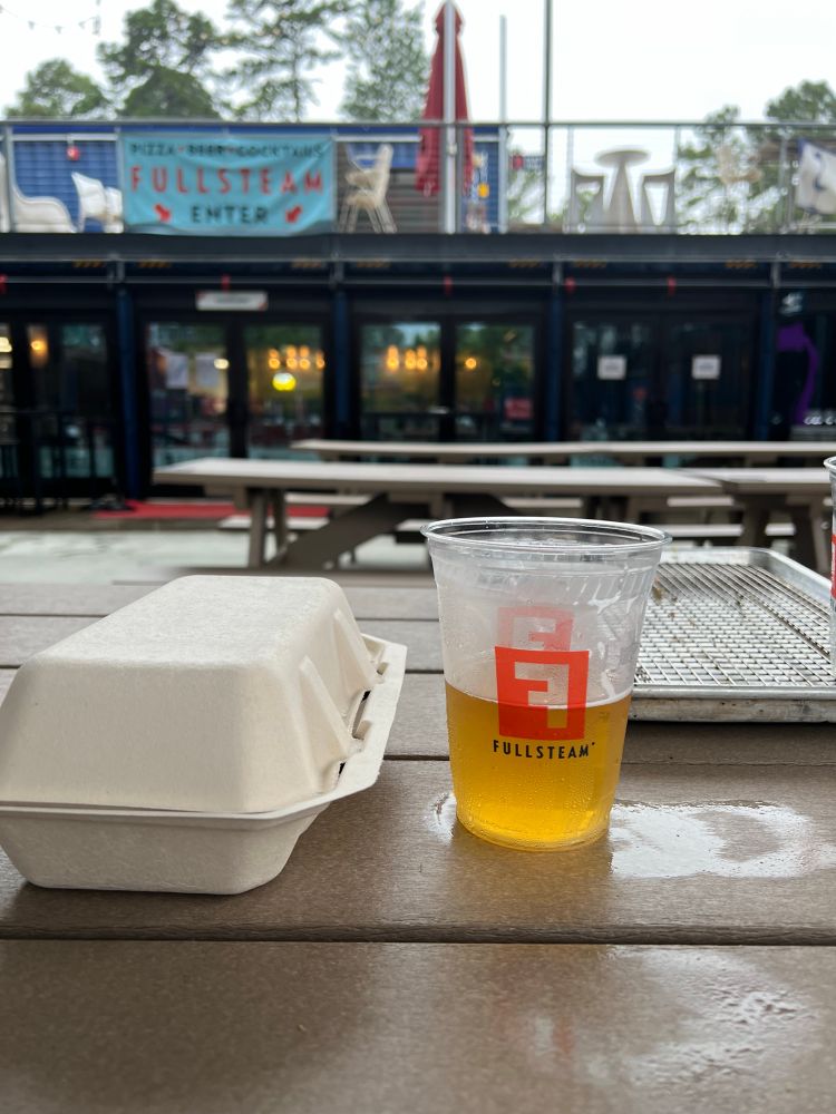Takeout box and plastic cup of beer on picnic table.  Raining in the background 
