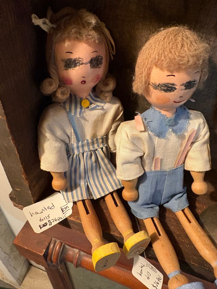 Two old time wooden dolls on a shelf. Their eyes are scratched out and they have a tag saying “haunted dolls $26 pair”