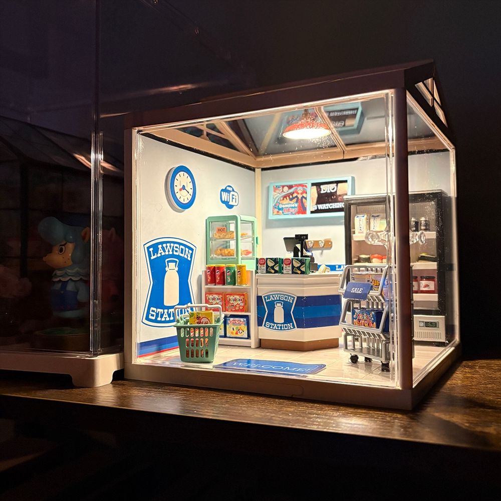 A miniature model of a Lawson convenience store on a bookshelf. It has a miniature counter and freezer filled with tiny food. There is a hanging light lighting up the miniature room. 