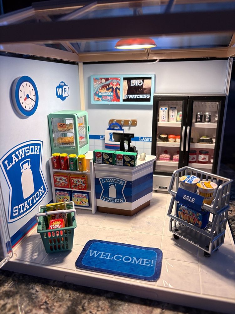 Close up of miniature Lawson convenience store. Details showing the small food on the shelves, counter, and in the freezer. There's a clock on the left wall as well as an ad for Dominos pizzer starring Hatsune Miku. There is also a picture of a one-eyed cat staring at you with the font 'Dio is watching' over him.