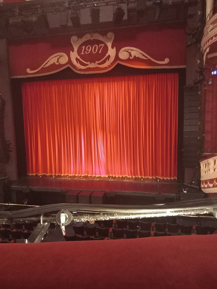 Large theatre stage with red curtains pulled closed over a dark brown stage. Some audience seats can be seen.