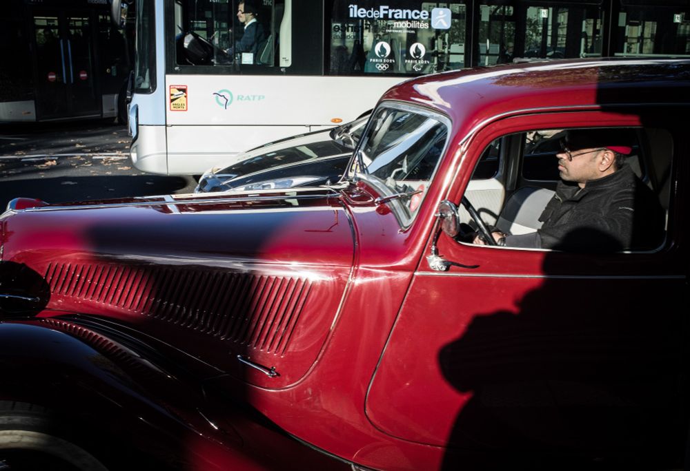 Une photo prise dans la rue à Paris.
Au premier plan une voiture ancienne, rouge. A l'arrière plan, un bus de la RATP.
Paris, octobre 2024.