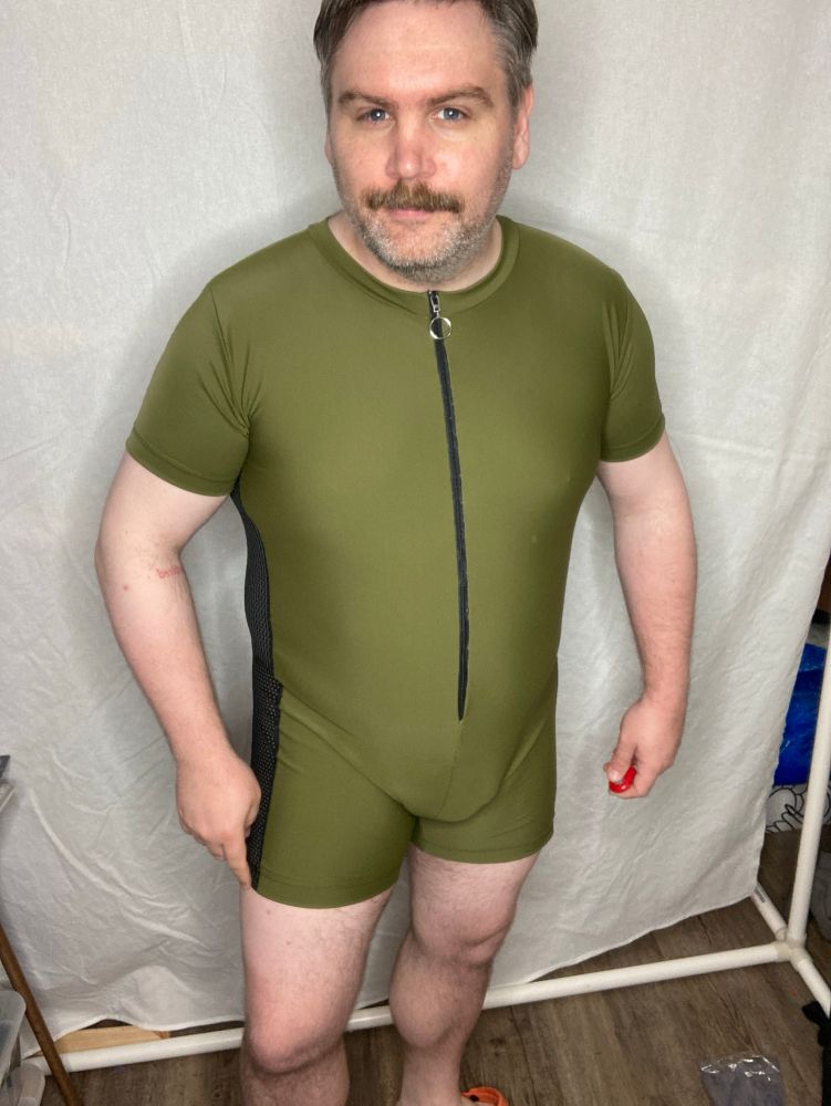 Me, white mustache 30s, way more gray than I realized wearing a drab olive green bodysuit with black mesh sides in front of a white background