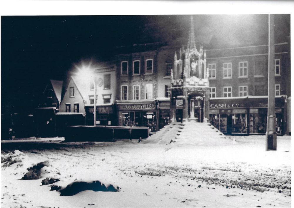 The Market Cross in snow Leighton Buzzard. The photo is black and white and taken with the shops behind. The cross is 5 sided and reached by a set of stone steps. 