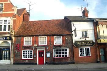 A picture of a pub with a red door in the centre, 2 windows either side, one a bay. Three windows upstairs. Squished between 2 buildings. A two storey extension added to one end. 