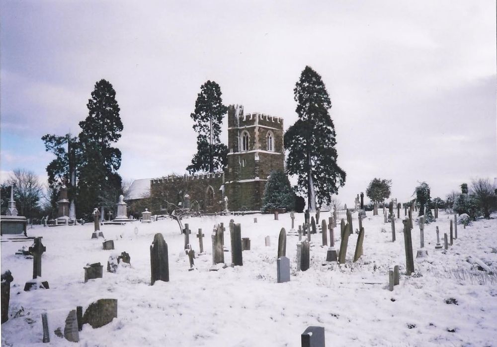 A stone church with a Norman style tower in the background, flanked by 3 large redwood trees. In the foreground gravestones in a churchyard covered in snow. 