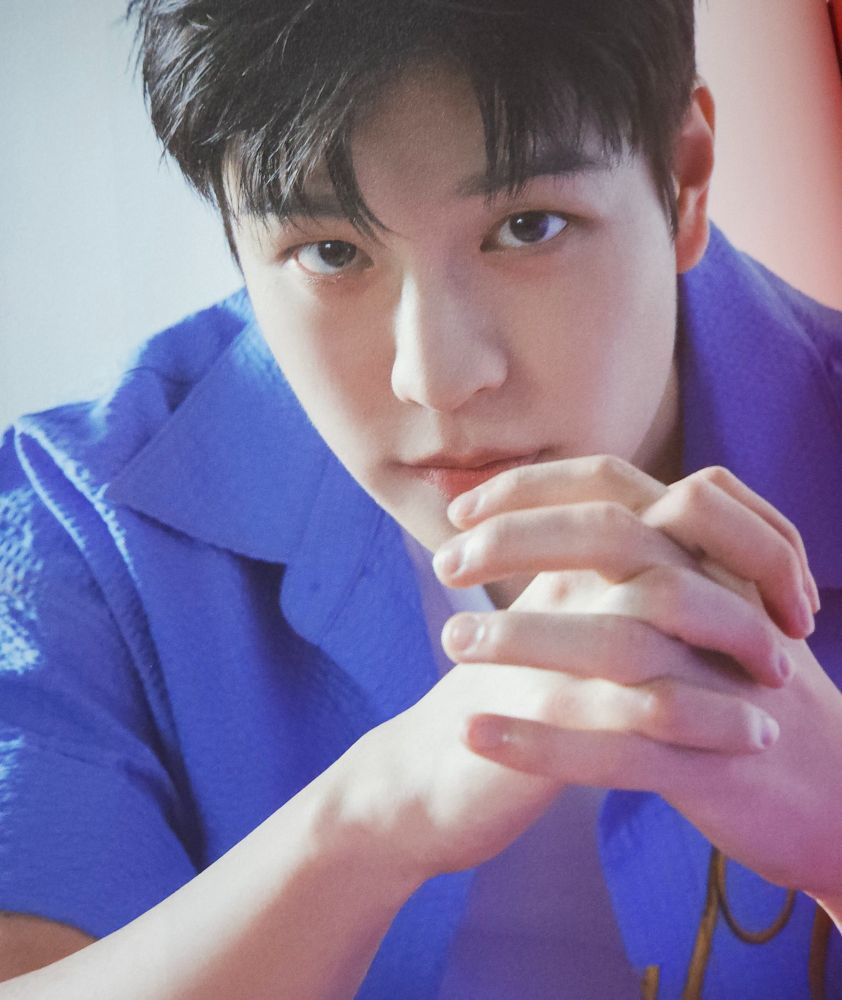 Seungmin is wearing a white blouse and a blue button-down shirt with gold letters. He is sitting down, with a white wall in the background and he is looking directly at the camera. His hands are crossed at the height of his chin. The angle of the photo is top down. 