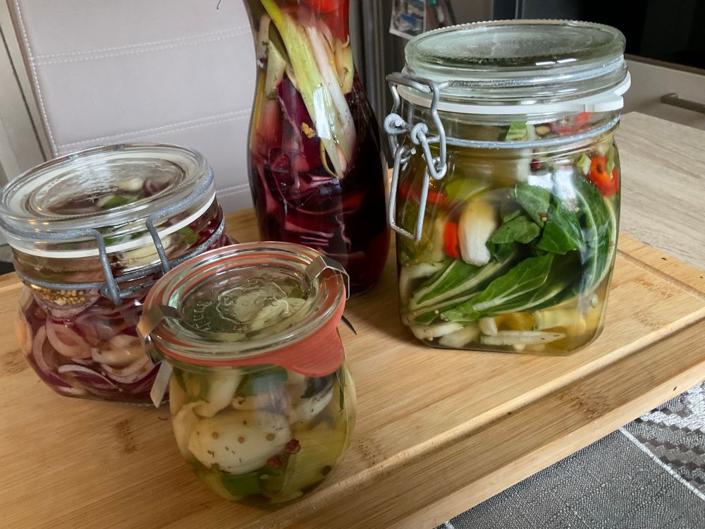 Auf einem Schneiderbrett befinden sich vier Einmachgläser mit Gemüse :Knoblauch,rote Zwiebel,Pak Choi,Rotkohl und Lauchzwiebel.Alle samt würden in einer Lösung aus Essig,Salz und Zucker sowie Wasser eingelegt.Außerdem wurde noch Chili,Pfeffer sowie Senfkörner dazu gegeben..Man kann alles dazu packen 