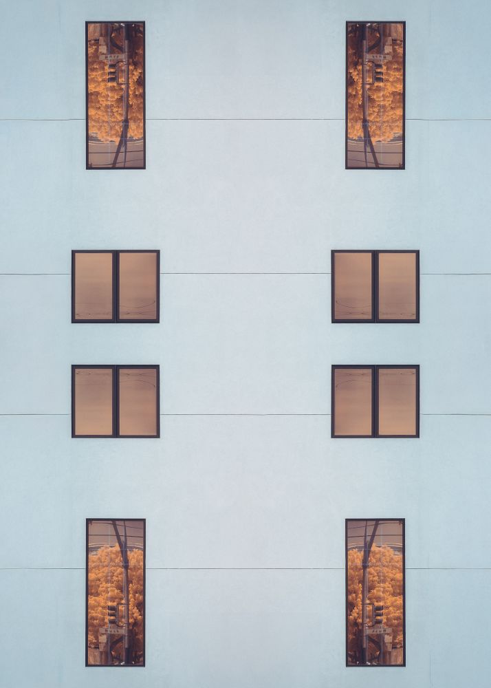 A mostly featureless bluish-gray wall with horizontal seams along it at regular intervals. Extremely plain, square windows with black frames are placed in the wall. Along the outside edges, tall windows have a reflection of a tree with orange leaves, a street sign, and a traffic light. The reflection is in a different orientation on each edge. Along the inside, a shorter pair of windows reflect a smooth progression of various shades of orange.