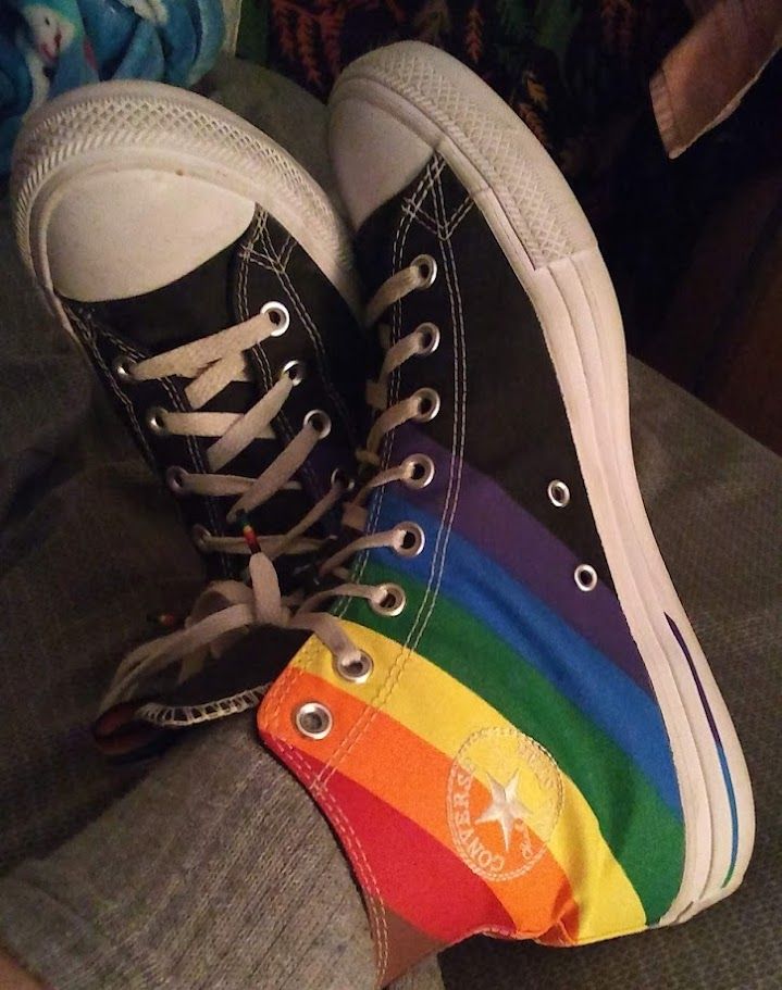 It says with no explanation, but nah, that leaves out y'all with vision impairments. all the love. *ahem*.. A 3 year old picture of my 2020 Pride Collection Chuck Taylor high top shoes with my feet propped up on my bed and crossed. The shoes have a white rubber outsole, black canvas with the Progress Pride (rainbow with brown and black) colors moving diagonally taking up most of the inner side of the shoe and wrapping around to the outer side. The typical "Chuck Patch" on the inner side is white embroidery, to place the branding, but not taking away from or covering up the colors.