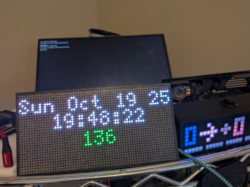a 64x32 HUB75 display a.k.a. Tidbyt screen bring driven with the correct date and time as well as the author's current blood glucose reading. Next to it, a Sugarpixel has a DNS lookup failure