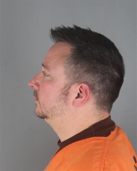 a large, thick-necked white guy with a middle-point haircut who has obviously just been crying stares to the side in a brown shirt underneath prison orange