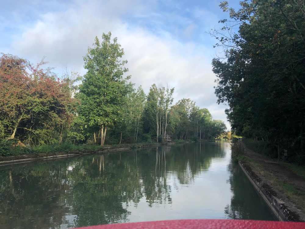 Morning view from Isobel’s roof hatch of the sunny tree lined canal under a blue sky with moody grey clouds.
