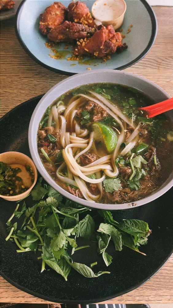 A bowl of short rib pho with mint and spicy chili sauce with Korean chicken wings in the background. 