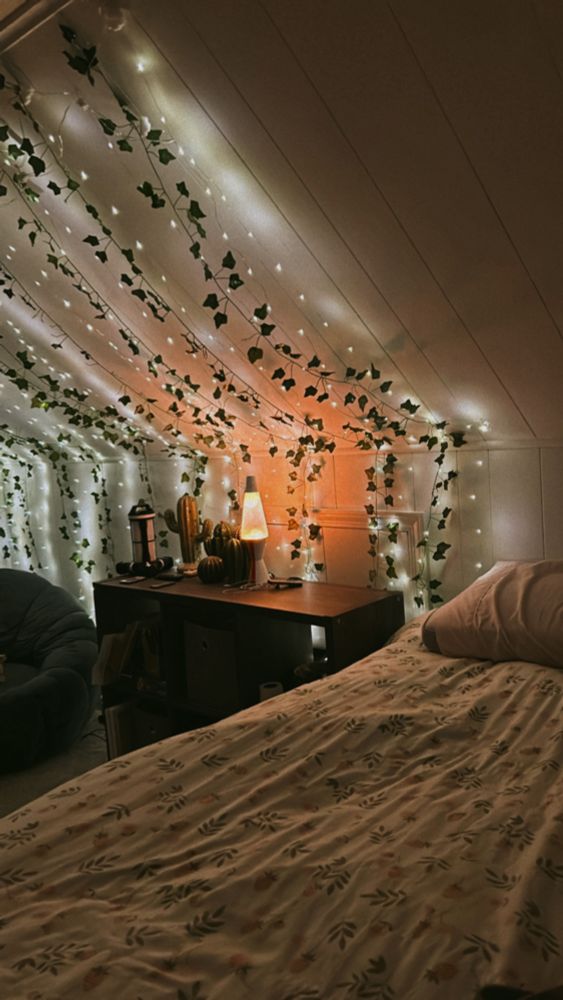 Photo of a spare bedroom with a slanted roof, showing half a bed, a decorated shelf unit and a small section of a puffy chair. There is a lava lamp on the shelf. Vines and string lights hang from the ceiling down the slant. 