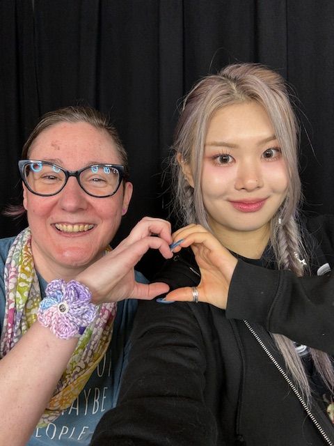 An White woman with brown hair and glasses making a hand heart with a younger Korean woman with pale blonde hair.