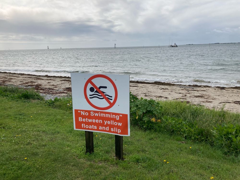 A sign at a beach reads “No swimming” in quotation marks, followed by other text which reads: between hello floats and slip. (Without quote marks on the latter text)