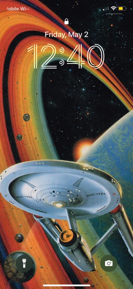 An iPhone Lock Screen featuring an illustration of The Enterprise, with a planet in the background that has big colorful rings around it. The illustration was originally the cover of a book, so there’s lots of empty space in the composition at the top where the title would go, allowing the time to be displayed without obscuring any of the major details. 