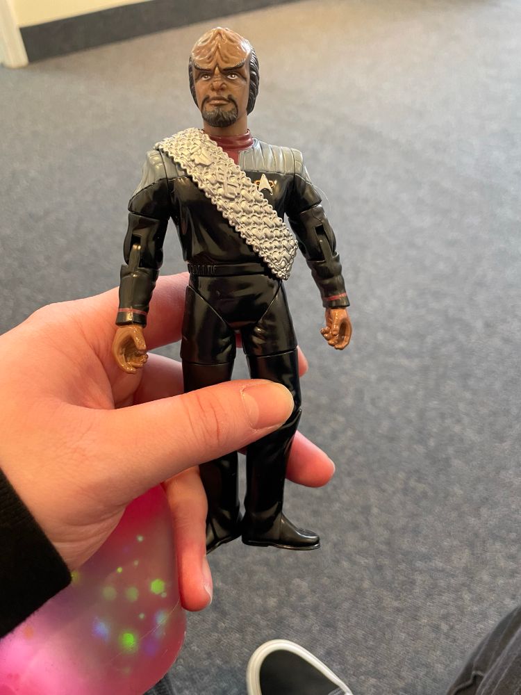 My hand holding a Worf action figure and a pink stress ball. The background is the ubiquitous dull grey carpet of a waiting room. 