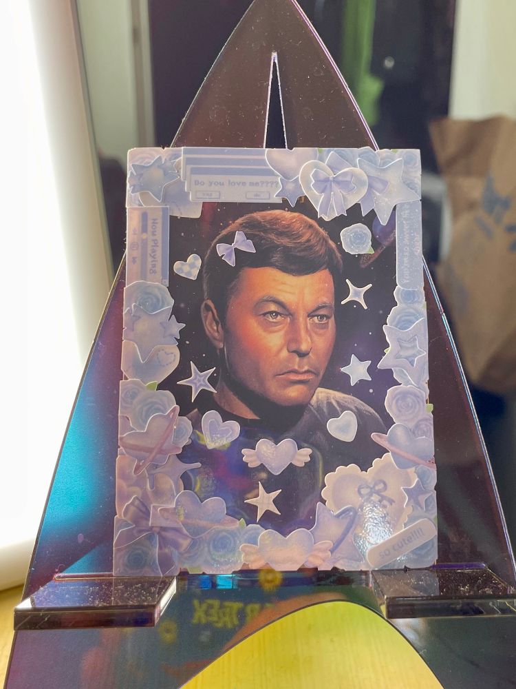 A trading card featuring an illustration of TOS-era Leonard McCoy. The card has been put into a clear card sleeve, which has a border on it made of stickers. The stickers are all cutesy and blue, mostly hearts, stars, bows, and flowers. 