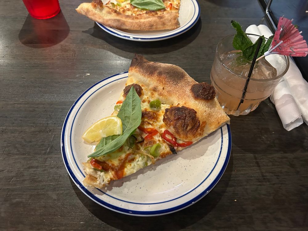 a slice of pizza on a plate and a cocktail. the pizza features several veggies, a large basil leaf, and a lemon slice. the cocktail is pink and features a sprig of mint and a pink umbrella. 