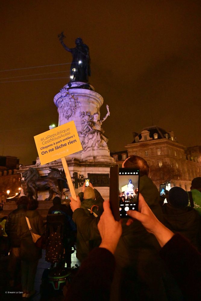 Au fond, la statue de la République illuminée. Au premier plan, une personne prend en photo une pancarte où il  est écrit "La République c'est nous aussi. Citoyens à part entière. On ne lâche rien !"