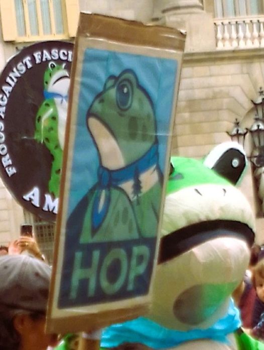 Close-up of picture in quoted post: in Barcelona, a person in frog costume, a round sign saying "frogs against fascism" and in front, a frog in profile over the word "HOP" 